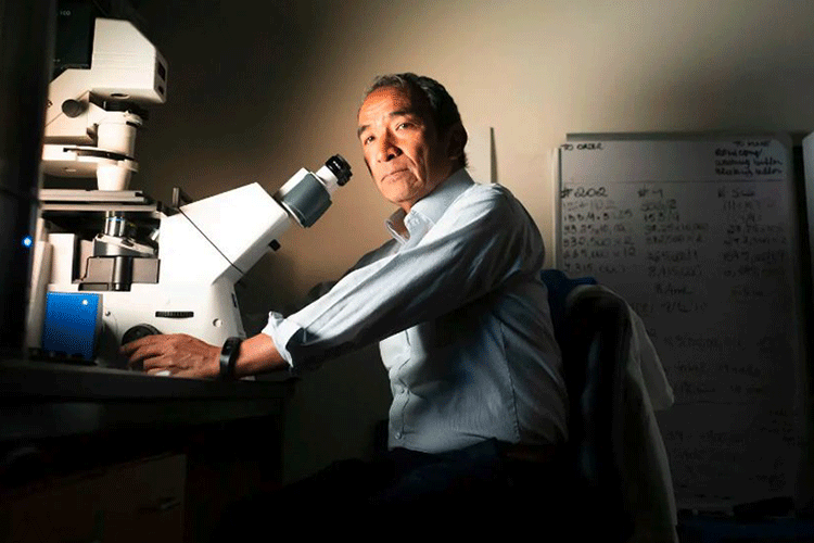 A photo of Lee Riley in a dark room. Riley is sitting in front of a microscope and is looking at the camera
