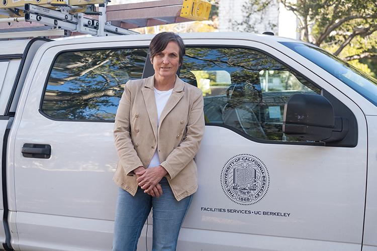 Diane Coppini, director of engineering technical sevices at UC Berkeley's Facilities Services, leans against a white Facilities Services vehicle. She is wearing a beige blazer and jeans and is looking at the camera with her hands folded at her waist.
