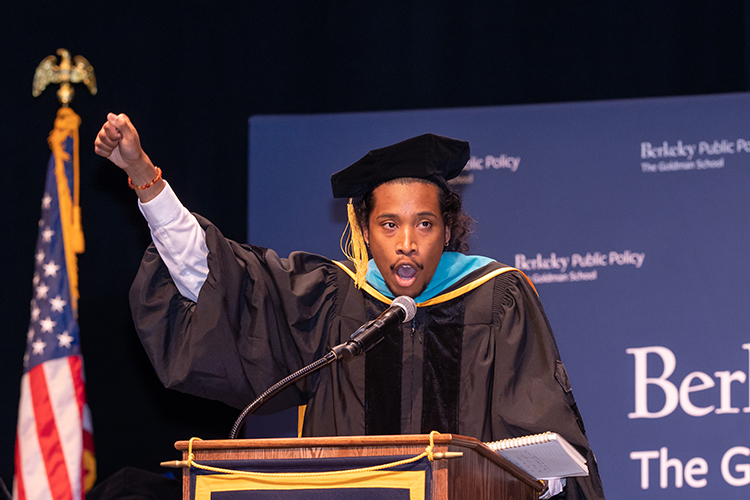 a person wearing a cap and gown holds fist in the air as he speaks at a podium