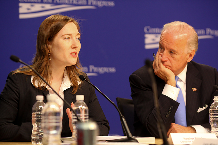 two people sit at a table with microphones while one of them talks to an audience outside of the frame