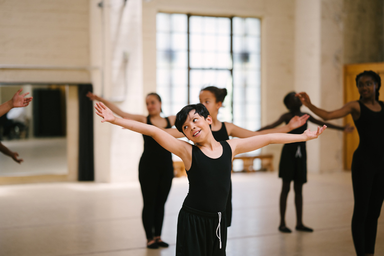 Several young dancers wearing a black leotards hold their arms out straight to either side as they practice in a well-lit gym