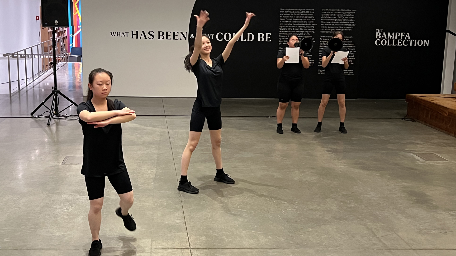 four dancers wearing black shirts, shorts and shoes perform a live dance in a museum gallery
