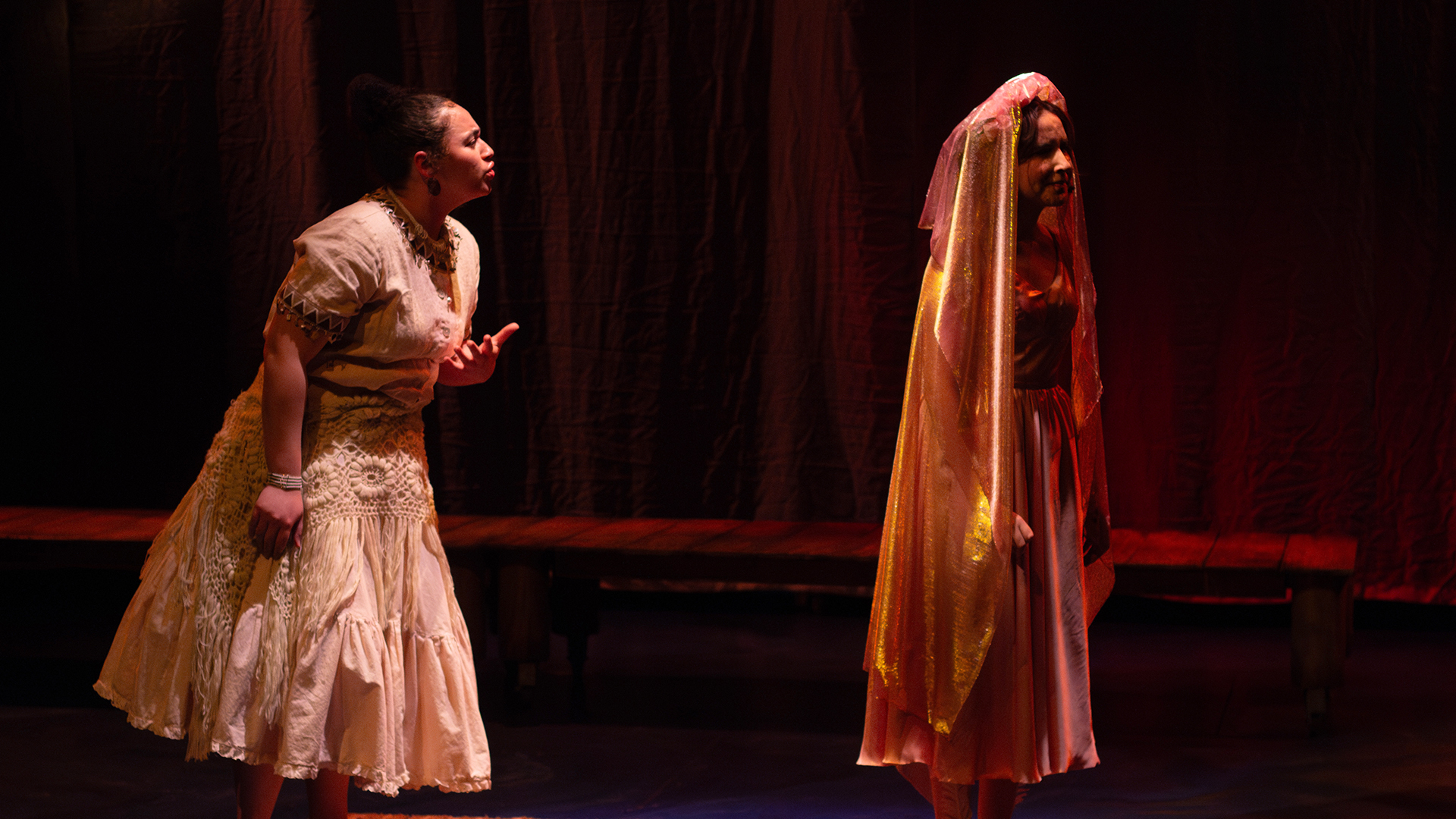 Two actors interact on stage, bathed in a reddish orange light. One actor, wearing a wedding dress and a veil, turns away from the other actor, who is facing her back and appears to be pleading with her.