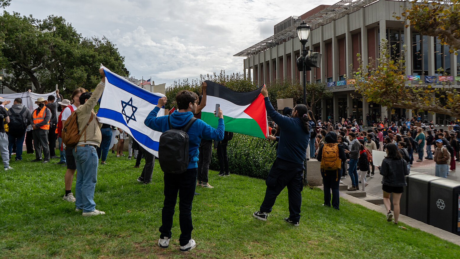 Two students hold an Israeli flag and two students hold up a Palestinian flag standing next to one another on the Berkeley campus with at least one hundred students on Sproul Plaza in the background