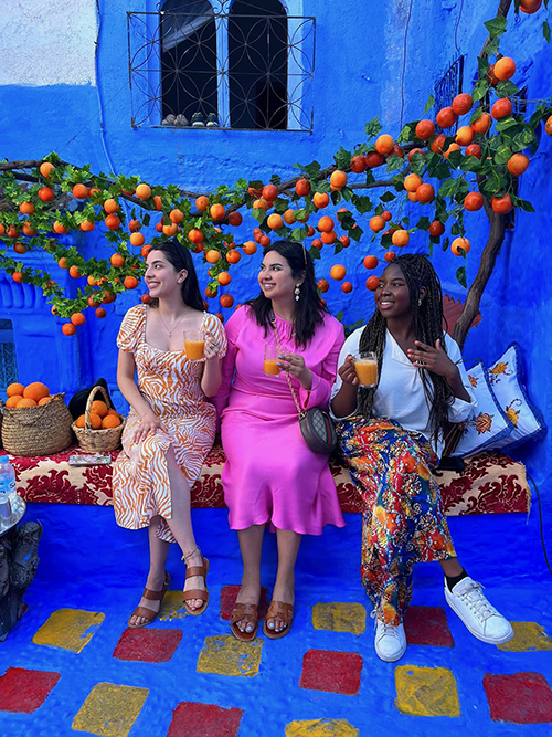 Daniella and two friends sit in front of a brilliant blue wall and ground with an orange tree growing behind them in Morocco. They are smiling and each holds a glass of orange juice.