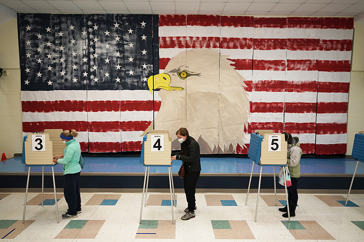 three people voting at booths with a giant American flag with an image of a bald eagle on the wall behind them